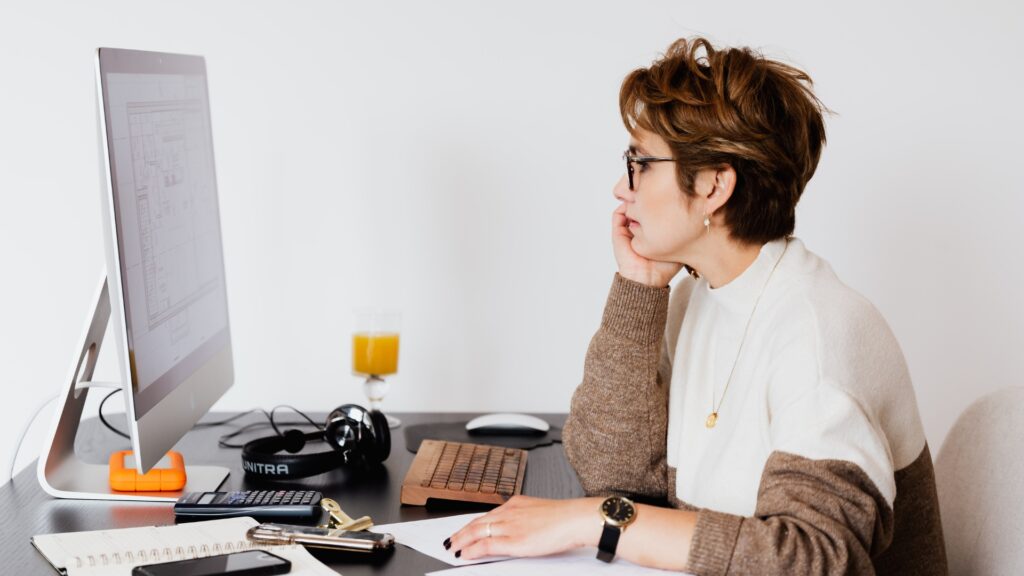 A side profile photo of a focused woman with short, curly brown hair and glasses, looking intently at a computer screen that displays what appears to be complex data or a spreadsheet. She rests her chin on her hand, surrounded by a cluttered desk with a calculator, notebook, phone, headphones, and a drink, embodying the stress of managing small business finances.