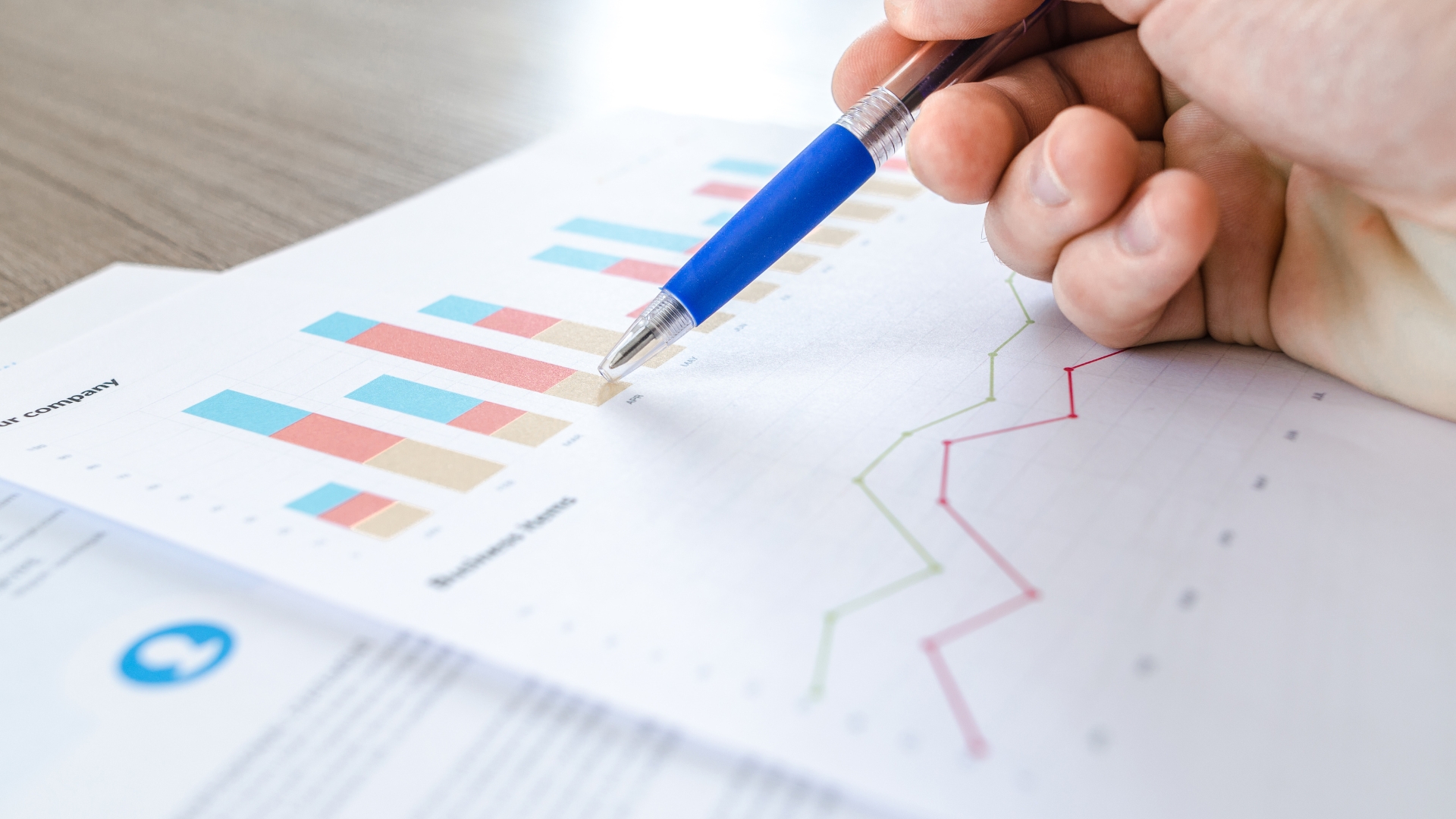 A close-up photo of a hand holding a blue pen, pointing to a colorful bar chart and line graph on a printed business report, symbolizing professional financial analysis and business growth.